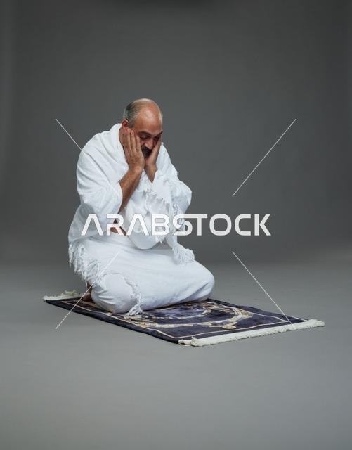 بورتريه لحاج كبير السن عربي سعودي خليجي بملابس الإحرام  يجلس على سجادة صلاه ويرفع يديه بالدعاء, اداء مناسك الحج والعمره , خلفيه رماديه