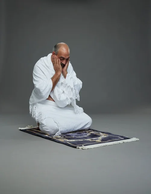 بورتريه لحاج كبير السن عربي سعودي خليجي بملابس الإحرام  يجلس على سجادة صلاه ويرفع يديه بالدعاء, اداء مناسك الحج والعمره , خلفيه رماديه