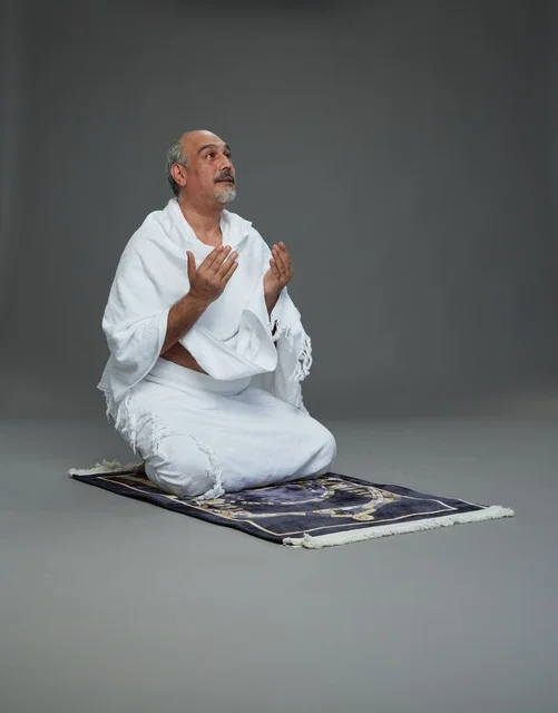 بورتريه لحاج كبير السن عربي سعودي خليجي بملابس الإحرام  يجلس على سجادة صلاه ويرفع يديه بالدعاء, اداء مناسك الحج والعمره , خلفيه رماديه