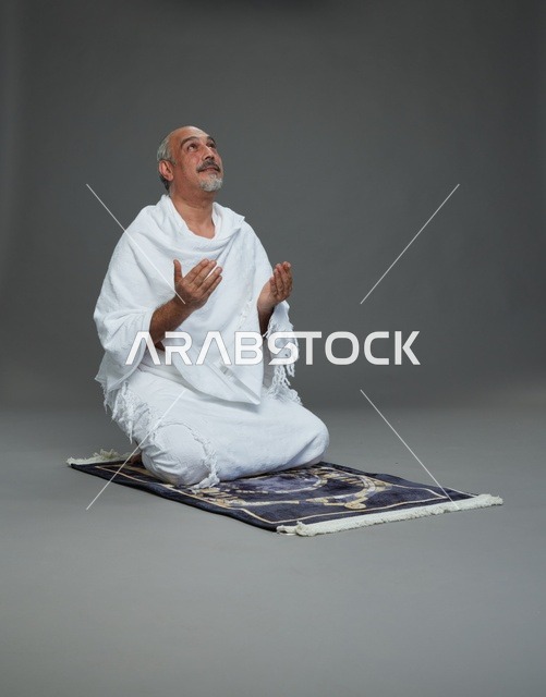 بورتريه لحاج كبير السن عربي سعودي خليجي بملابس الإحرام  يجلس على سجادة صلاه ويرفع يديه بالدعاء, اداء مناسك الحج والعمره , خلفيه رماديه