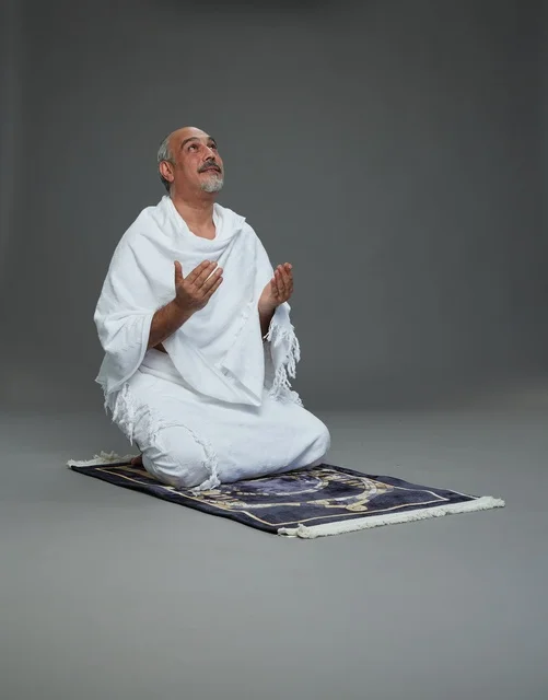 بورتريه لحاج كبير السن عربي سعودي خليجي بملابس الإحرام  يجلس على سجادة صلاه ويرفع يديه بالدعاء, اداء مناسك الحج والعمره , خلفيه رماديه