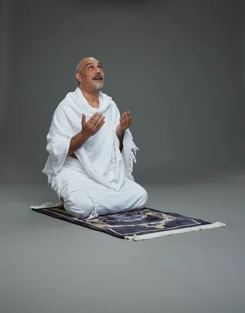 بورتريه لحاج كبير السن عربي سعودي خليجي بملابس الإحرام  يجلس على سجادة صلاه ويرفع يديه بالدعاء, اداء مناسك الحج والعمره , خلفيه رماديه