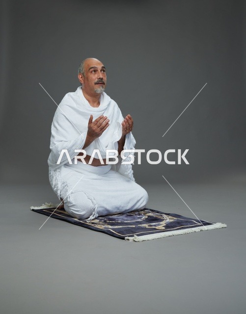 بورتريه لحاج كبير السن عربي سعودي خليجي بملابس الإحرام  يجلس على سجادة صلاه ويرفع يديه بالدعاء, اداء مناسك الحج والعمره , خلفيه رماديه