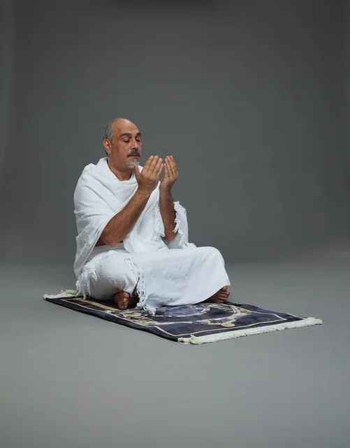 A portrait of an elderly Arab Saudi Gulf pilgrim in Ihram clothing sitting on a prayer rug, raising his hands in prayer, performing the rituals of Hajj and Umrah, with a gray background.