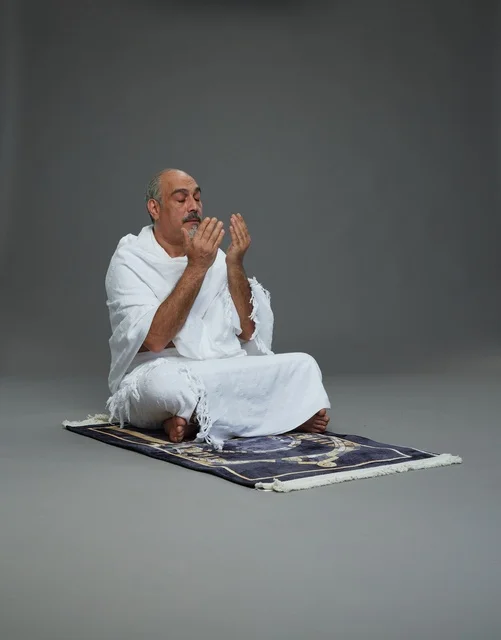 بورتريه لحاج كبير السن عربي سعودي خليجي بملابس الإحرام  يجلس على سجادة صلاه ويرفع يديه بالدعاء, اداء مناسك الحج والعمره , خلفيه رماديه