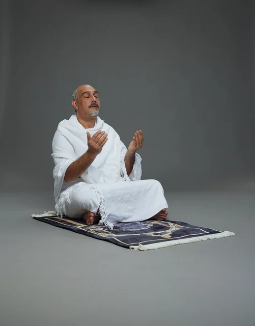 بورتريه لحاج كبير السن عربي سعودي خليجي بملابس الإحرام  يجلس على سجادة صلاه ويرفع يديه بالدعاء, اداء مناسك الحج والعمره , خلفيه رماديه