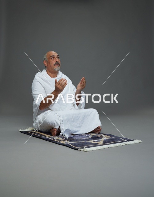 بورتريه لحاج كبير السن عربي سعودي خليجي بملابس الإحرام  يجلس على سجادة صلاه ويرفع يديه بالدعاء, اداء مناسك الحج والعمره , خلفيه رماديه
