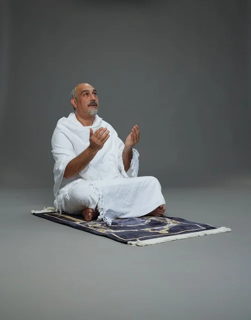 بورتريه لحاج كبير السن عربي سعودي خليجي بملابس الإحرام  يجلس على سجادة صلاه ويرفع يديه بالدعاء, اداء مناسك الحج والعمره , خلفيه رماديه