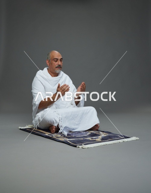بورتريه لحاج كبير السن عربي سعودي خليجي بملابس الإحرام  يجلس على سجادة صلاه ويرفع يديه بالدعاء, اداء مناسك الحج والعمره , خلفيه رماديه