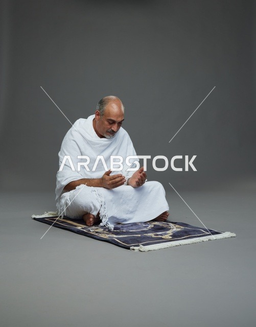 بورتريه لحاج كبير السن عربي سعودي خليجي بملابس الإحرام  يجلس على سجادة صلاه ويرفع يديه بالدعاء, اداء مناسك الحج والعمره , خلفيه رماديه