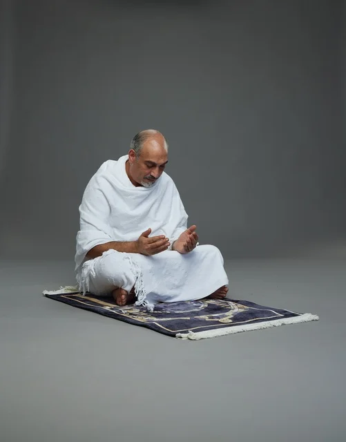 بورتريه لحاج كبير السن عربي سعودي خليجي بملابس الإحرام  يجلس على سجادة صلاه ويرفع يديه بالدعاء, اداء مناسك الحج والعمره , خلفيه رماديه