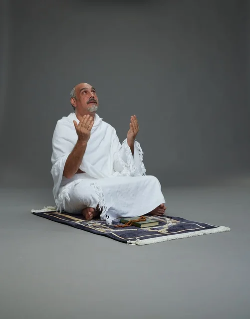 بورتريه لحاج كبير السن عربي سعودي خليجي بملابس الإحرام  يجلس على سجادة صلاه ويرفع يديه بالدعاء, اداء مناسك الحج والعمره , خلفيه رماديه