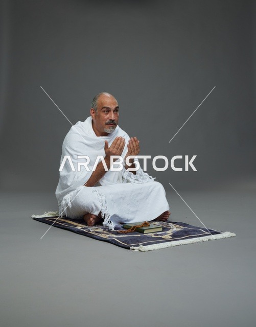 بورتريه لحاج كبير السن عربي سعودي خليجي بملابس الإحرام  يجلس على سجادة صلاه ويرفع يديه بالدعاء, اداء مناسك الحج والعمره , خلفيه رماديه
