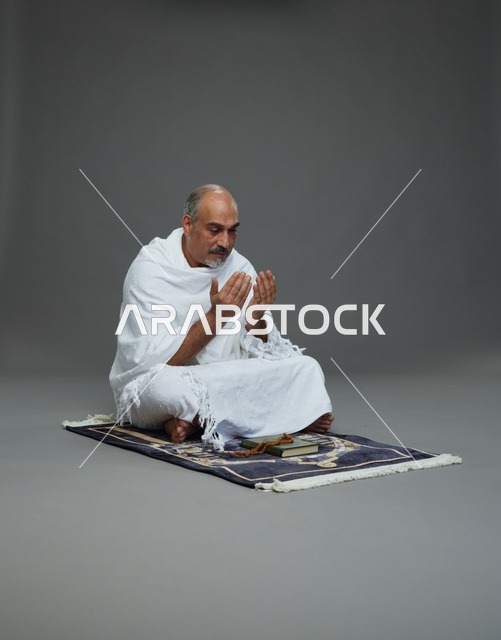 بورتريه لحاج كبير السن عربي سعودي خليجي بملابس الإحرام  يجلس على سجادة صلاه ويرفع يديه بالدعاء, اداء مناسك الحج والعمره , خلفيه رماديه