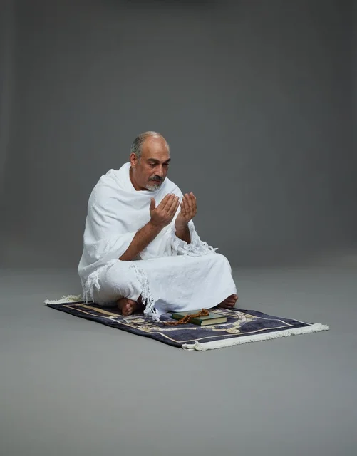 A portrait of an elderly Arab Saudi Gulf pilgrim in Ihram clothing sitting on a prayer rug, raising his hands in prayer, performing the rituals of Hajj and Umrah, with a gray background.