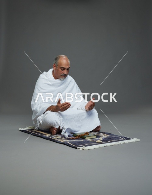 بورتريه لحاج كبير السن عربي سعودي خليجي بملابس الإحرام  يجلس على سجادة صلاه ويرفع يديه بالدعاء, اداء مناسك الحج والعمره , خلفيه رماديه