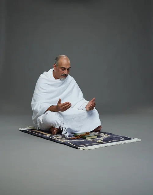 بورتريه لحاج كبير السن عربي سعودي خليجي بملابس الإحرام  يجلس على سجادة صلاه ويرفع يديه بالدعاء, اداء مناسك الحج والعمره , خلفيه رماديه