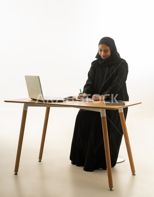 Saudi Woman Writing at Office Desk with Laptop and Notebook