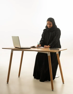 Saudi Woman Writing at Office Desk with Laptop and Notebook
