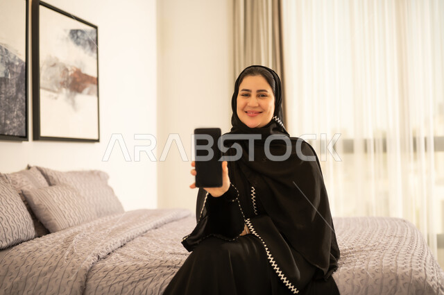 امرأة سعودية بالعباءة تحمل هاتفاً ذكياً في غرفة النوم