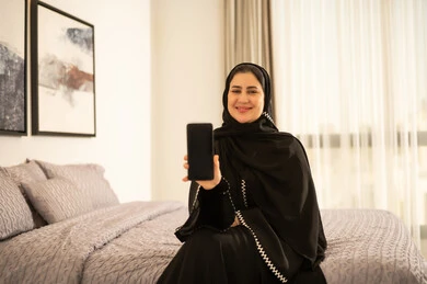 امرأة سعودية بالعباءة تحمل هاتفاً ذكياً في غرفة النوم