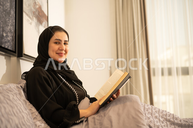 امرأة سعودية مبتسمة تقرأ كتاباً في غرفة نوم عصرية