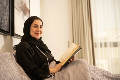 امرأة سعودية مبتسمة تقرأ كتاباً في غرفة نوم عصرية
