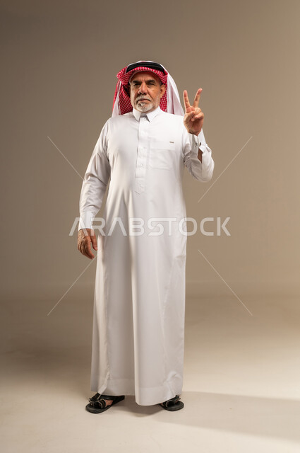 رجل سعودي مسن يؤدي إيماءة النصر في استوديو محايد