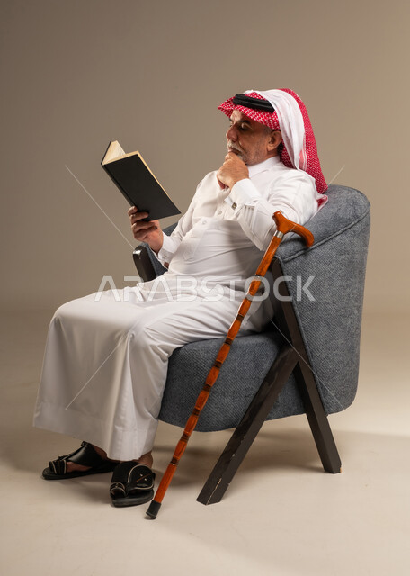 رجل سعودي مسن يقرأ كتاباً على كرسي مريح مع عصا مشي