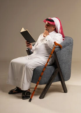رجل سعودي مسن يقرأ كتاباً على كرسي مريح مع عصا مشي