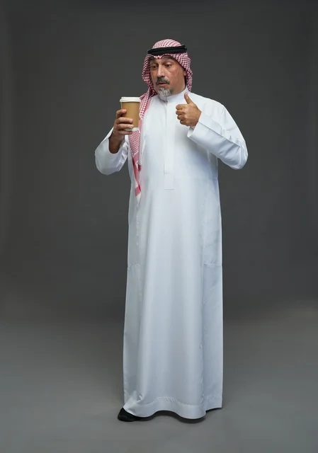 رجل  عربي سعودي خليجي مسن يرتدي الزي الثوب الابيض والشماغ ويحمل فنجان قهوة,  ايماءات واشارات مختلفه , كامل الجسم, خلفيه رماديه