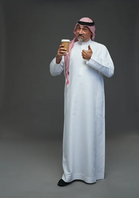 رجل  عربي سعودي خليجي مسن يرتدي الزي الثوب الابيض والشماغ ويحمل فنجان قهوة,  ايماءات واشارات مختلفه , كامل الجسم, خلفيه رماديه