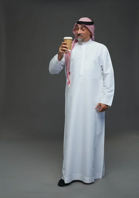 An elderly Arab Saudi Gulf man wearing a white thobe and a shemagh, holding a cup of coffee, with various gestures and signs, full body, gray background.
