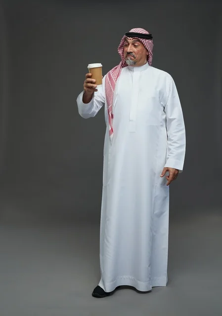 رجل  عربي سعودي خليجي مسن يرتدي الزي الثوب الابيض والشماغ ويحمل فنجان قهوة,  ايماءات واشارات مختلفه , كامل الجسم, خلفيه رماديه