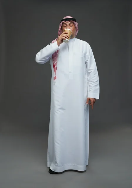 رجل  عربي سعودي خليجي مسن يرتدي الزي الثوب الابيض والشماغ ويحمل فنجان قهوة,  ايماءات واشارات مختلفه , كامل الجسم, خلفيه رماديه