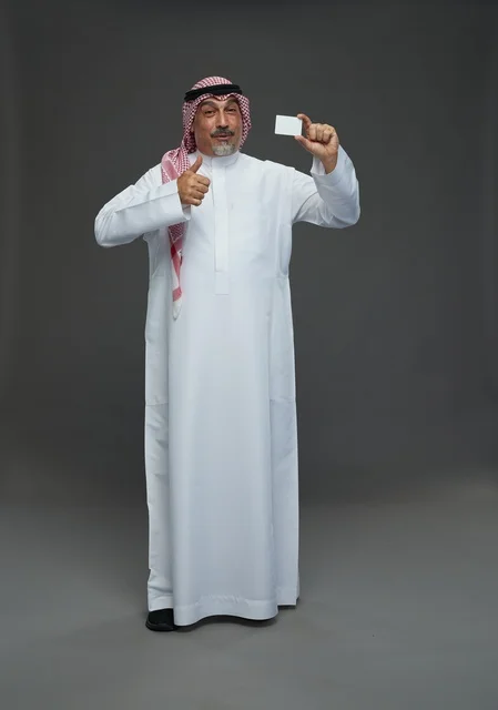رجل  عربي سعودي خليجي مسن يرتدي الزي الثوب الابيض والشماغ ويمسك بطاقه بيضاء فارغه,  ايماءات واشارات مختلفه , كامل الجسم, خلفيه رماديه