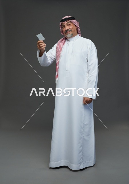 رجل  عربي سعودي خليجي مسن يرتدي الزي الثوب الابيض والشماغ ويمسك بطاقه بيضاء فارغه,  ايماءات واشارات مختلفه , كامل الجسم, خلفيه رماديه
