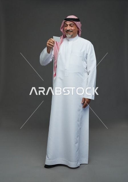 رجل  عربي سعودي خليجي مسن يرتدي الزي الثوب الابيض والشماغ ويمسك بطاقه بيضاء فارغه,  ايماءات واشارات مختلفه , كامل الجسم, خلفيه رماديه