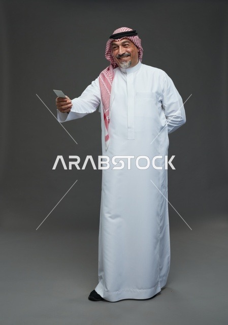 رجل  عربي سعودي خليجي مسن يرتدي الزي الثوب الابيض والشماغ ويمسك بطاقه بيضاء فارغه,  ايماءات واشارات مختلفه , كامل الجسم, خلفيه رماديه