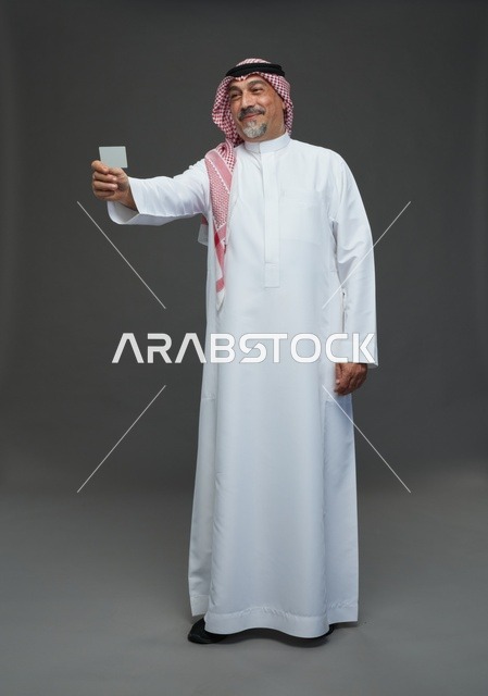 رجل  عربي سعودي خليجي مسن يرتدي الزي الثوب الابيض والشماغ ويمسك بطاقه بيضاء فارغه,  ايماءات واشارات مختلفه , كامل الجسم, خلفيه رماديه