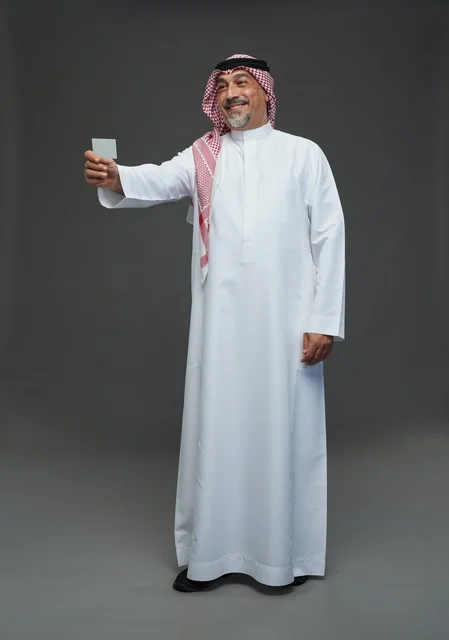 An elderly Arab Saudi Gulf man wearing a white thobe and a shemagh, holding a blank white card, with various gestures and signs, full body, gray background.