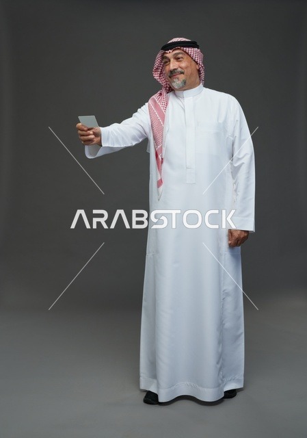 رجل  عربي سعودي خليجي مسن يرتدي الزي الثوب الابيض والشماغ ويمسك بطاقه بيضاء فارغه,  ايماءات واشارات مختلفه , كامل الجسم, خلفيه رماديه