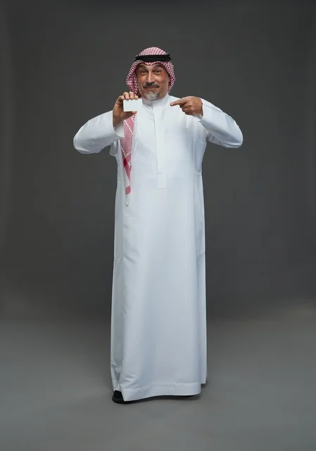 رجل  عربي سعودي خليجي مسن يرتدي الزي الثوب الابيض والشماغ ويمسك بطاقه بيضاء فارغه,  ايماءات واشارات مختلفه , كامل الجسم, خلفيه رماديه