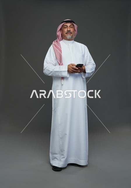 رجل  عربي سعودي خليجي مسن يرتدي الزي الثوب الابيض والشماغ ويمسك الهاتف المحمول,  ايماءات واشارات مختلفه , كامل الجسم, خلفيه رماديه