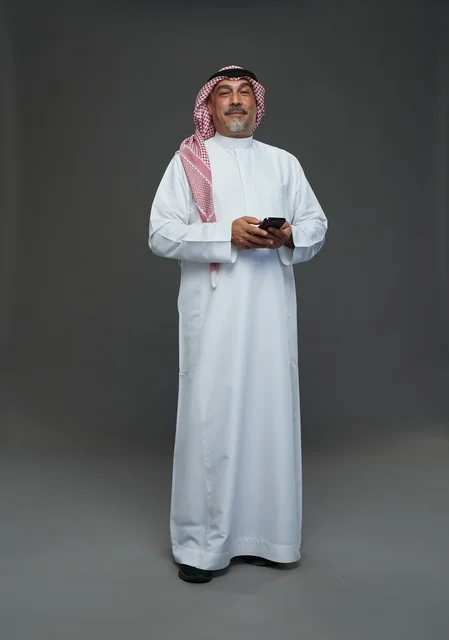 رجل  عربي سعودي خليجي مسن يرتدي الزي الثوب الابيض والشماغ ويمسك الهاتف المحمول,  ايماءات واشارات مختلفه , كامل الجسم, خلفيه رماديه