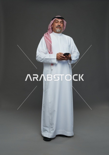 رجل  عربي سعودي خليجي مسن يرتدي الزي الثوب الابيض والشماغ ويمسك الهاتف المحمول,  ايماءات واشارات مختلفه , كامل الجسم, خلفيه رماديه