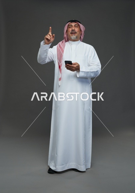 رجل  عربي سعودي خليجي مسن يرتدي الزي الثوب الابيض والشماغ ويمسك الهاتف المحمول,  ايماءات واشارات مختلفه , كامل الجسم, خلفيه رماديه
