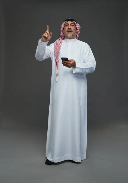 رجل  عربي سعودي خليجي مسن يرتدي الزي الثوب الابيض والشماغ ويمسك الهاتف المحمول,  ايماءات واشارات مختلفه , كامل الجسم, خلفيه رماديه
