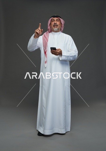 رجل  عربي سعودي خليجي مسن يرتدي الزي الثوب الابيض والشماغ ويمسك الهاتف المحمول,  ايماءات واشارات مختلفه , كامل الجسم, خلفيه رماديه