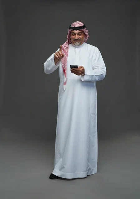 رجل  عربي سعودي خليجي مسن يرتدي الزي الثوب الابيض والشماغ ويمسك الهاتف المحمول,  ايماءات واشارات مختلفه , كامل الجسم, خلفيه رماديه